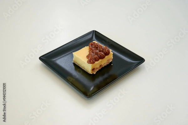 Obraz A slice of cheesecake with nuts on top. It is on a black plate. The plate is on a white table