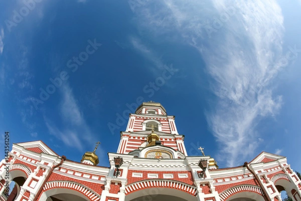 Obraz A vibrant red and white striped bell tower of a Russian Orthodox church with golden domes, set against a bright blue sky with wispy clouds. Architectural beauty.
