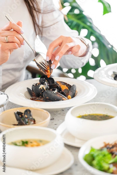 Obraz A close-up of a woman's hand expertly using a fork to select mussels from a plate, surrounded by bowls of vibrant dishes in a bright, modern dining setting.
