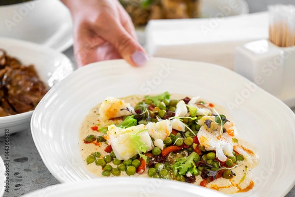 Obraz A hand presents a beautifully arranged plate featuring grilled fish with green peas and microgreens, creating a colorful and appetizing dish in a sophisticated dining environment.