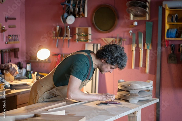 Fototapeta Professional woodworker verifying project details on mobile phone in joinery workshop. Examining customer requests, handmade woodworking, business administration, quality control, deadline management
