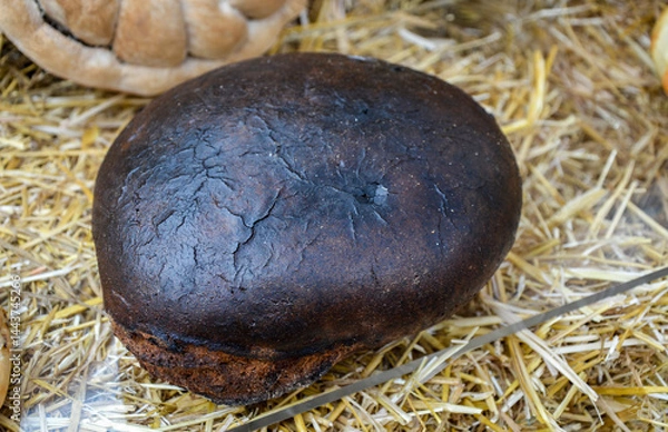 Obraz Dark, round loaf of bread with burnt crust rests on straw. Its cracked surface hints at high-temperature baking, showcasing rustic and artisanal style in a simple, natural setting