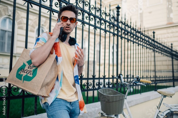 Fototapeta Young man with headphones and reusable shopping bag is having a phone call next to his bicycle