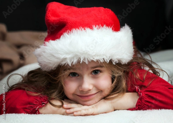 Obraz Girl with blonde hair in disguise of Santa Claus in a white-fur cap with a beard resting on his hands