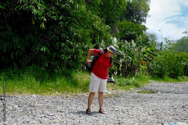 Fototapeta A woman is standing on a rocky path, adjusting her backpack, surrounded by green plant