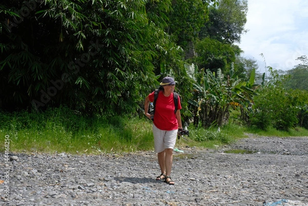Fototapeta Side view: A woman walks along on a rocky path surrounded by green trees under a bright; clear sky