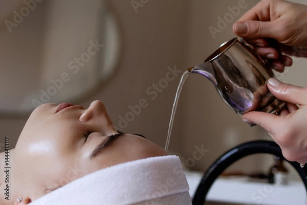 Obraz A woman is receiving a Japanese head spa treatment at a beauty salon. The therapist gently pours water, liquid onto her forehead to enhance relaxation of a scalp. Atmosphere promotes stress relief