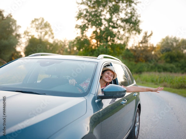 Fototapeta Traveler woman in red ethnic dress and hat drives car through scenic countryside of Croatia in summer. Surrounded by lush fields and forests, she enjoys peaceful and adventurous road trip