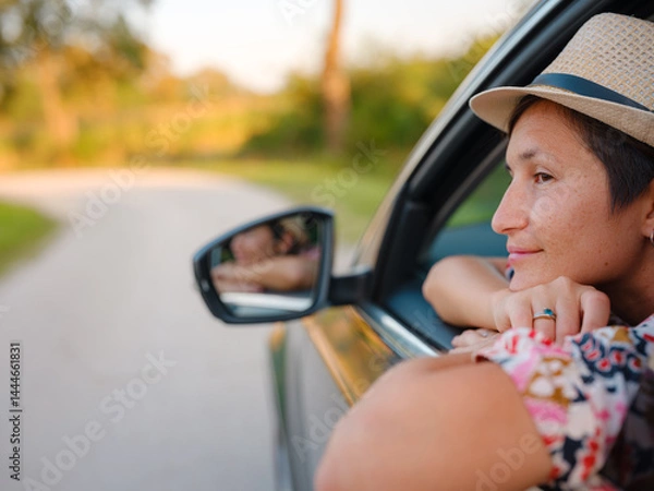Fototapeta Traveler woman in red ethnic dress and hat drives car through scenic countryside of Croatia in summer. Surrounded by lush fields and forests, she enjoys peaceful and adventurous road trip