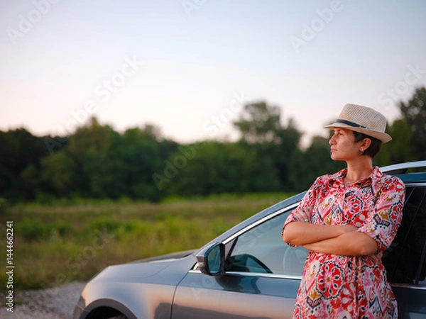 Fototapeta Traveler woman in red ethnic dress and hat drives car through scenic countryside of Croatia in summer. Surrounded by lush fields and forests, she enjoys peaceful and adventurous road trip