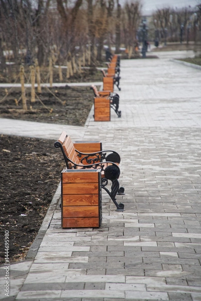 Fototapeta Row of wooden benches and matching planters along a paved walkway in a spring city park, ideal for illustrating urban landscaping, public seating, and recreational spaces. Photo