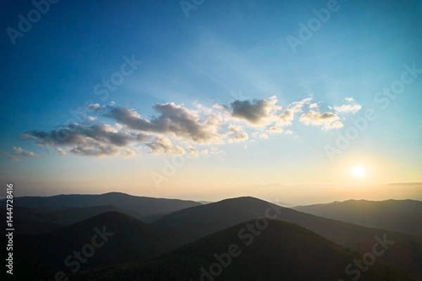 Obraz Serene sunset over rolling hills and mountains. Sky is gradient of blue and orange, with scattered clouds catching sun's rays. Landscape below cloaked in shadows, creating peaceful and tranquil scene.