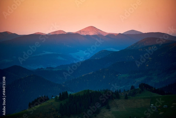 Obraz Sunset over mountain ridges with warm, orange-pink sky. Peaks highlighted with soft light, creating gradient of shadows across landscape. Small houses nestled among rolling hills.