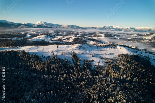 Obraz Aerial view reveals stunning winter landscape with snow-covered hills and dense forests. Houses nestled among trees, while background features majestic range of mountains under clear blue sky.