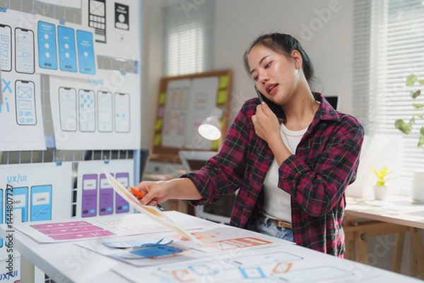 Obraz Female web designer reviewing mobile application wireframes while discussing project details on a phone call in a stylish, modern office environment, showcasing creativity and innovation