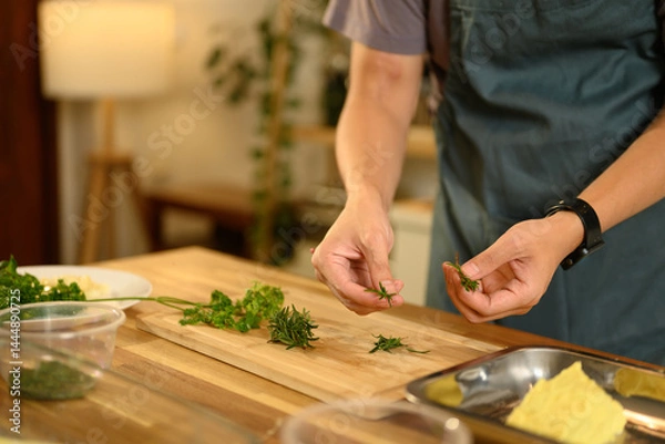 Fototapeta Man preparing fresh herbs on wooden cutting board for making compound butter in a kitchen