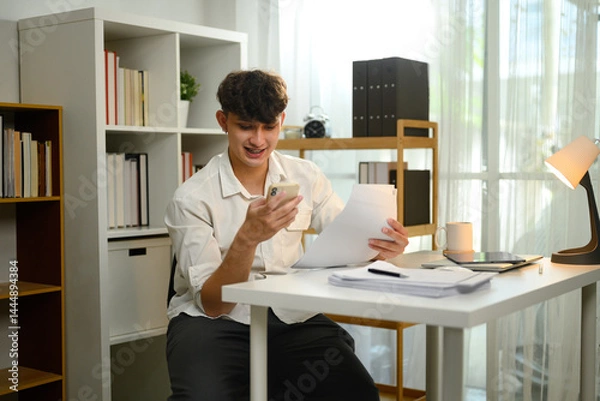 Fototapeta Smiling man using smartphone while reviewing documents in a home office