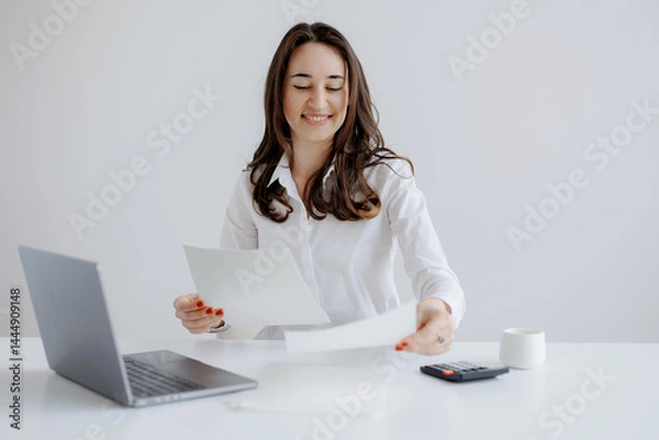 Fototapeta Young woman reviewing documents at a white desk with a laptop and calculator during a daytime office setting