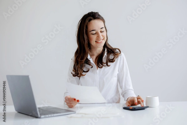 Fototapeta Woman working at a desk with laptop, calculator, and documents in a bright, minimalist workspace during the day