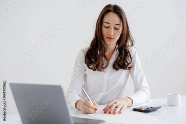 Fototapeta Young woman working diligently at a desk while taking notes and using a laptop in a bright, minimalist office setting