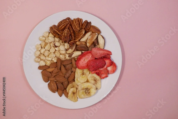 Fototapeta A variety of nuts and dried fruits on a plate on pink background. Hazelnuts, pecans, brazil nuts, almonds, dried strawberries and bananas. The concept of nutrition, diet, healthy eating and sports
