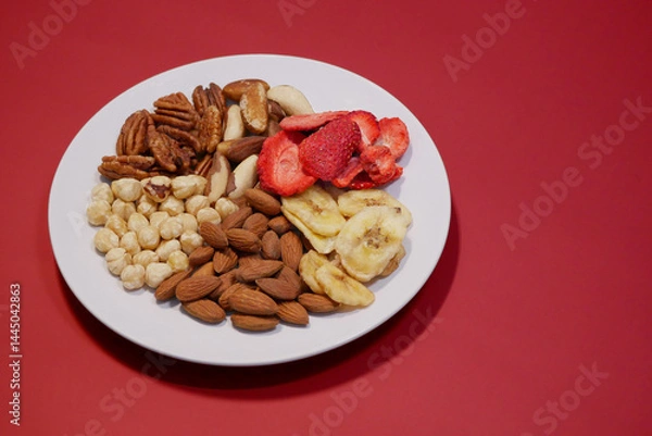 Fototapeta A variety of nuts and dried fruits on a plate on red background. Hazelnuts, pecans, brazil nuts, almonds, dried strawberries and bananas. The concept of nutrition, diet, healthy eating and sports