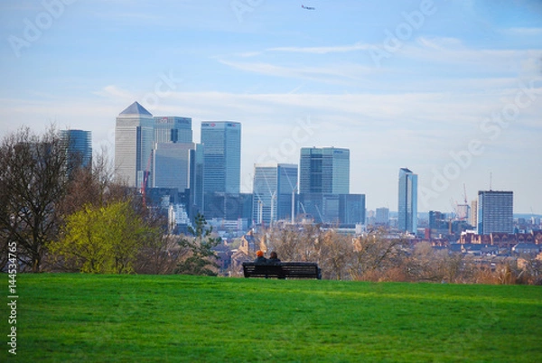 Fototapeta Couple sitting down on a bench on hill looking at the London Canary Wharf Skyline in Greenwich