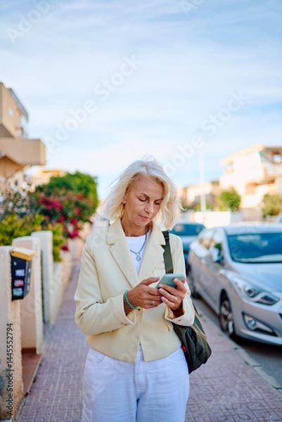 Fototapeta Woman walking on a sunny street checking her phone in a coastal town