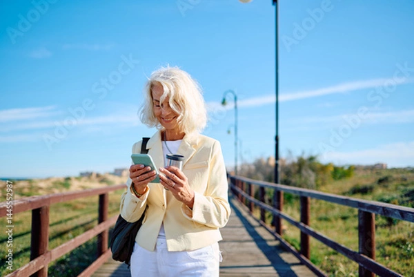 Fototapeta Senior woman enjoying a walk by the seaside while checking her phone