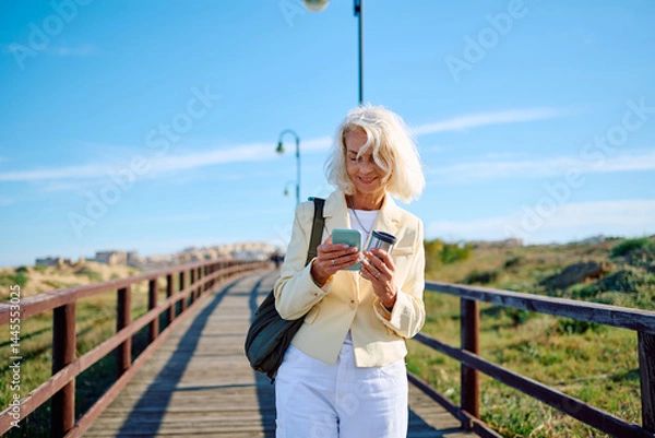 Fototapeta Woman checking smartphone while walking on wooden pier in sunny weather