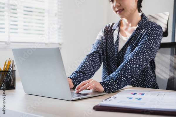 Fototapeta Businesswoman sitting at desk on couch in workplace or at home working on laptop and analyzing data on charts and graphs and writing on papers to make business plan and strategies for company, 