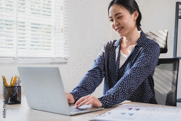 Fototapeta Businesswoman sitting at desk on couch in workplace or at home working on laptop and analyzing data on charts and graphs and writing on papers to make business plan and strategies for company, 