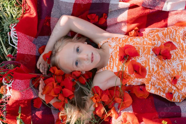 Obraz Smiling child girl on a picnic blanket among wildflowers, enjoying a sunny summer day in nature.