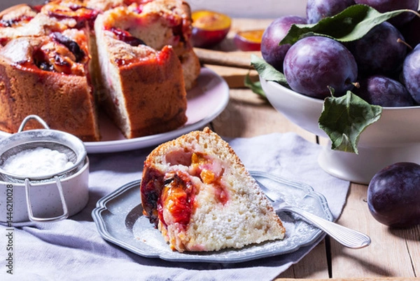 Fototapeta Plum pie with filling and sugar crust, sprinkled with powdered sugar and plums on a wooden table.