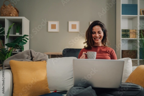Fototapeta Smiling woman drinking coffee and using laptop on sofa at home