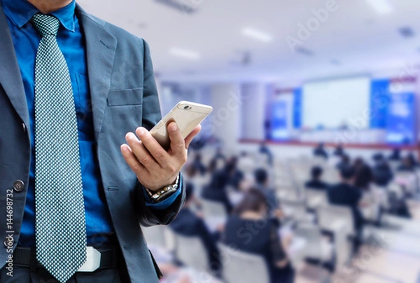 Fototapeta businessman using the mobile phone, blurred of conference hall or seminar room with attendee background.