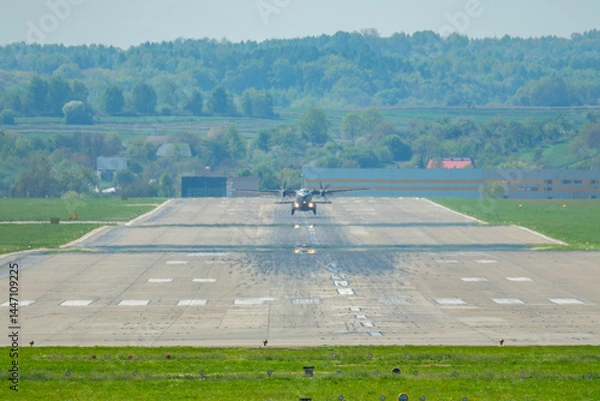 Fototapeta Military aircraft approaches runway during a clear day in a rural airfield surrounded by lush greenery and distant hills