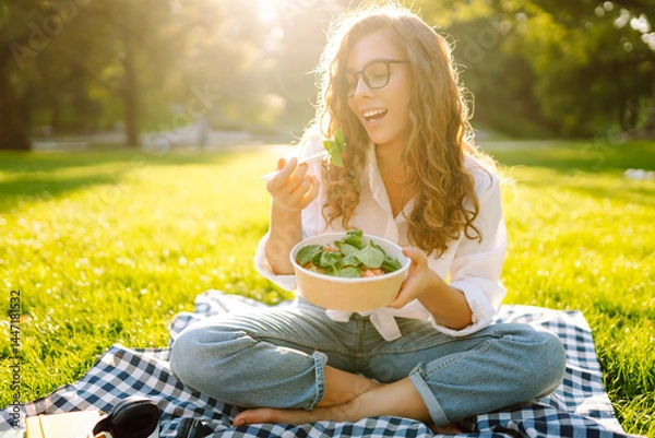 Fototapeta Happy woman in glasses sits on a mat in a park and eats a green salad. Young woman enjoys a snack in a park at sunset. Healthy eating, relaxation concept.