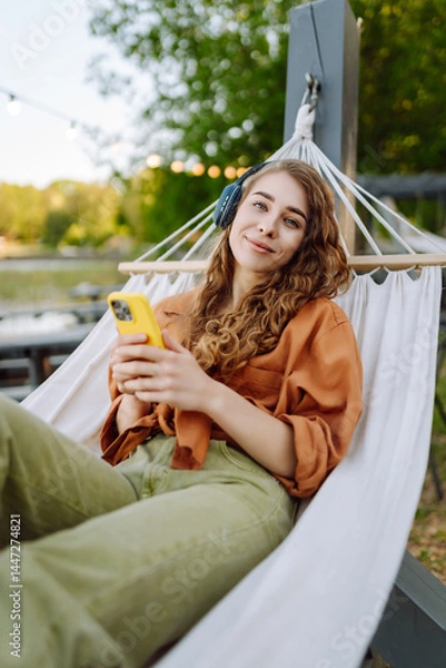 Fototapeta Young woman with curly hair listens to music on headphones while relaxing in a hammock. Cheerful woman smiling while using her phone in a sunny park. Leisure, blogging concept.