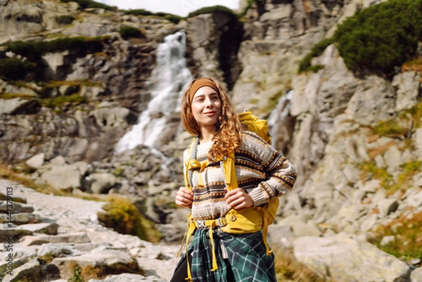 Fototapeta Young female traveler with a colorful backpack walking along a hiking trail with a mountain waterfall in the background. Beautiful woman enjoying nature and feeling freedom. Hiking concept.