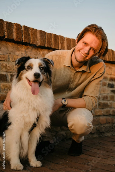Obraz Smiling young man in beige casual outfit kneels beside his happy Australian Shepherd dog during golden hour, with a rustic stone wall and warm sunset tones in the background.