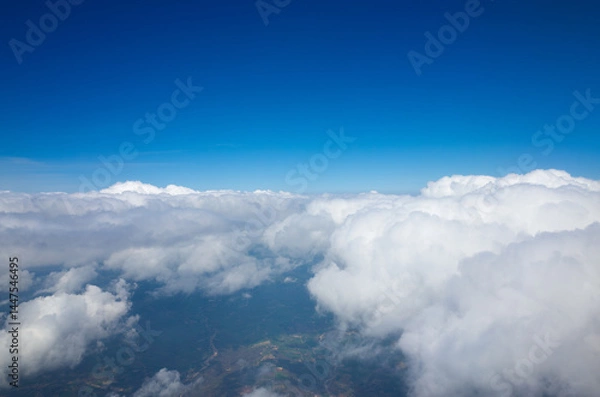 Fototapeta Aerial view from above a dense layer of clouds. The sky is a vibrant blue, contrasting with the fluffy white clouds below. Earth's surface are visible through breaks in the cloud cover