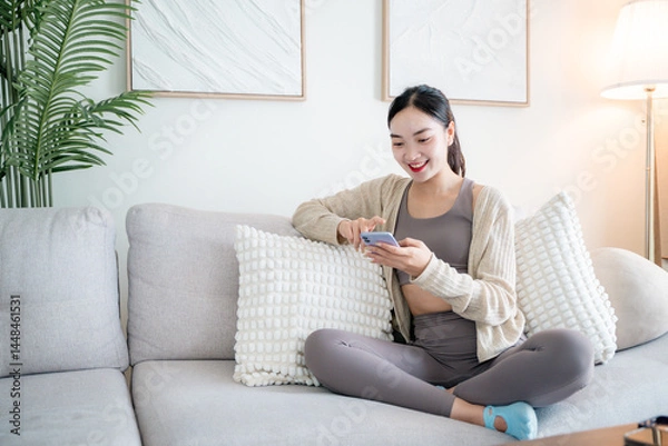 Fototapeta Smiling woman using mobile phone while sitting on sofa in living room