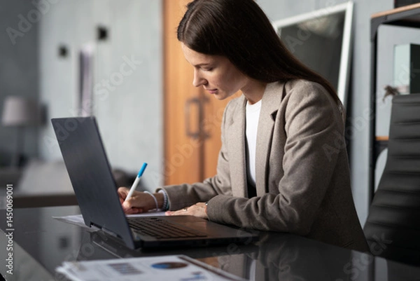 Fototapeta Young woman sitting at desk in office, working on her laptop and writing notes documents home office