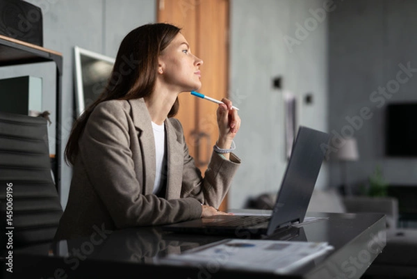 Fototapeta Young woman sitting at office desk, holding a pen to her chin in deep thought. laptop is open in front of her
