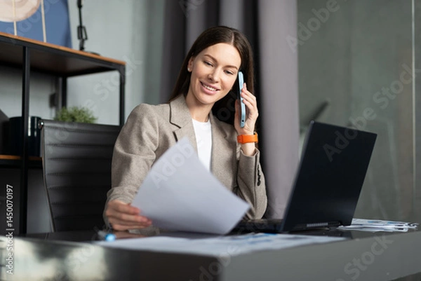 Fototapeta Business female talking on phone at desk and holding a document.