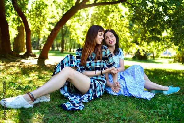 Fototapeta Two ladies resting on the grass in nature and talking to each other with smiles against the background of the spring park.