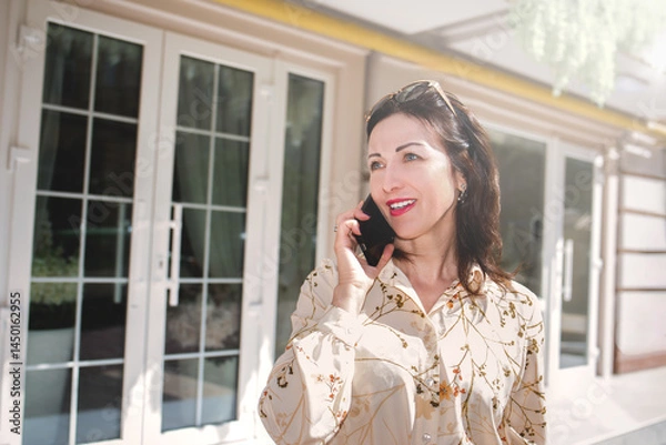 Fototapeta A portrait of a smiling woman wearing sunglasses, talking on the phone on the background of white windows of a house.