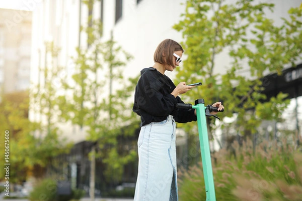 Obraz Stylish young woman with smartphone scans QR code for electric scooter rental. Girl using app on phone to unlock dockless transport on street. Urban style riding ecological vehicle. Modern technology