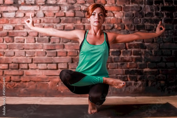 Fototapeta Young slender athletic girl in a turquoise tank top and dark leggings doing yoga against the background of a brick wall